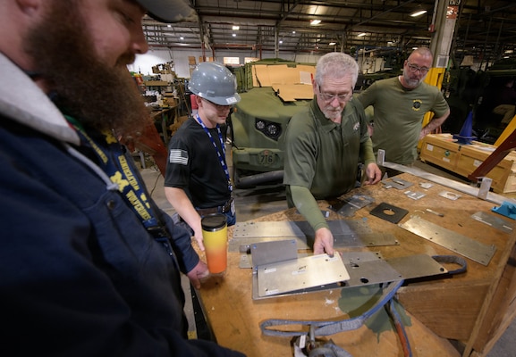 An older man in a green shirt demonstrates an assembly process with a metal plate to three colleagues gathered around a workbench in a workshop.