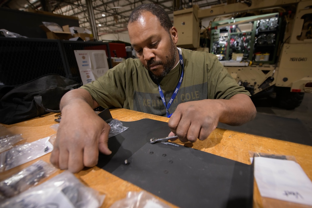 A man in a workshop carefully uses a small tool to fasten a metal component onto a black part, with a vehicle in the background.