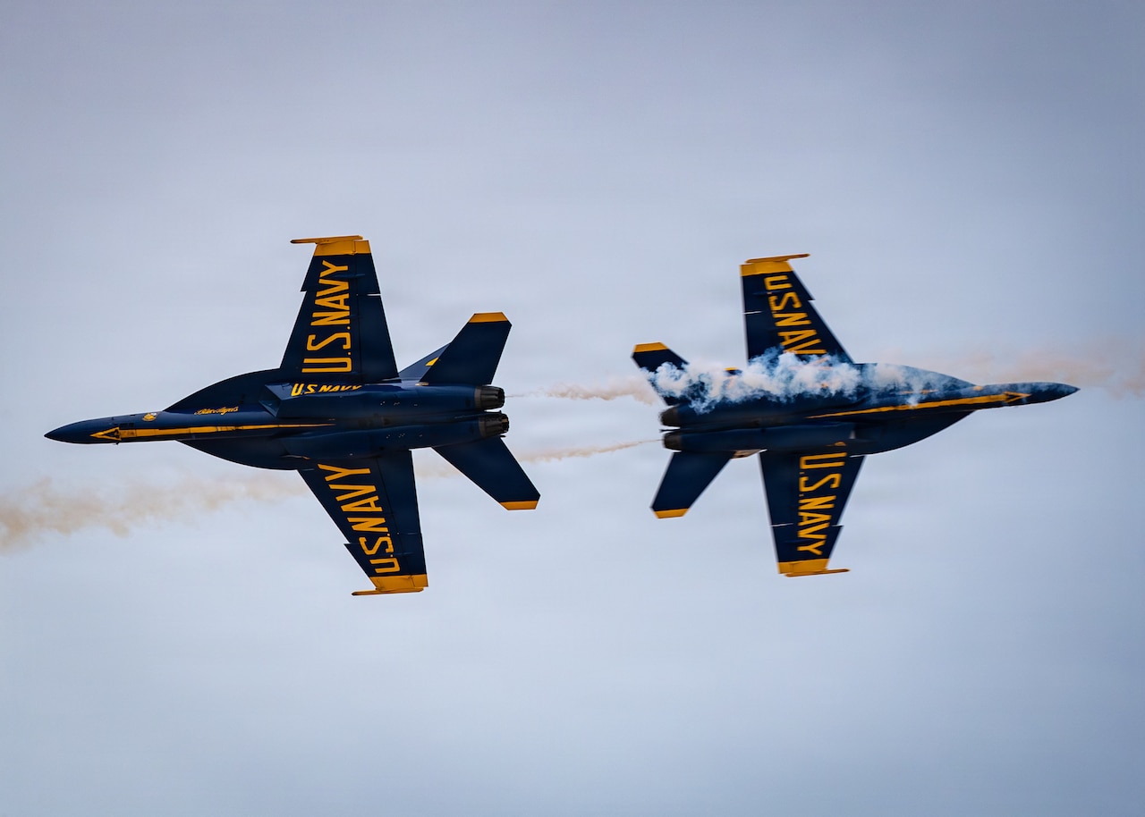 Two blue and yellow military show aircraft fly away from each other. Smoke billows from their exhaust areas.