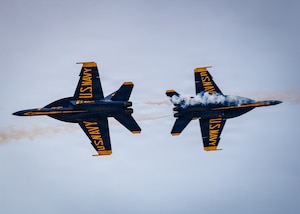 Two blue and yellow military show aircraft fly away from each other. Smoke billows from their exhaust areas.