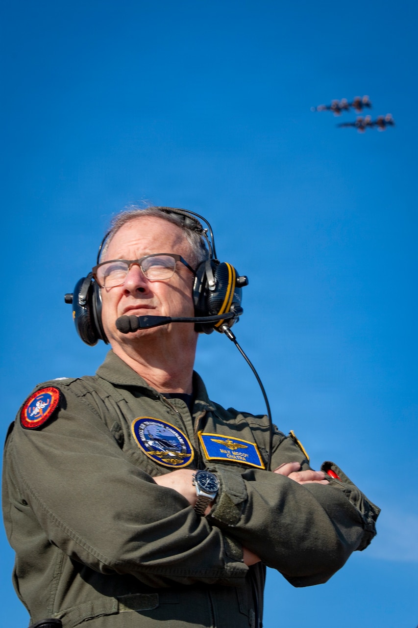 A man in a military flight suit looks to his right, as two military aircraft are seen in the distance above.