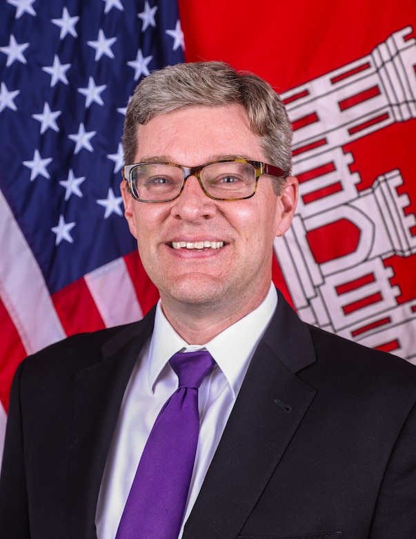 A man with glasses sits in front of the American and U.S. Army Corps of Engineers Flag, He is wearing a dark suit coat with a white collard shirt and a purple tie.