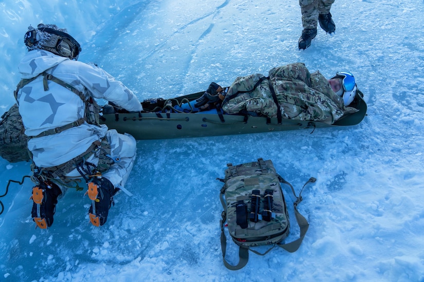 A U.S. airman assists a service member in a military stretcher on a snowy and icy surface as a backpack and binoculars rest nearby.