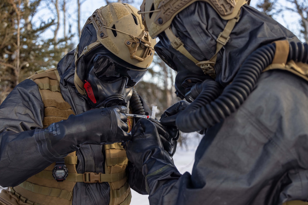 Two Marines in hazmat and tactical gear use a syringe while standing in a wooded area.
