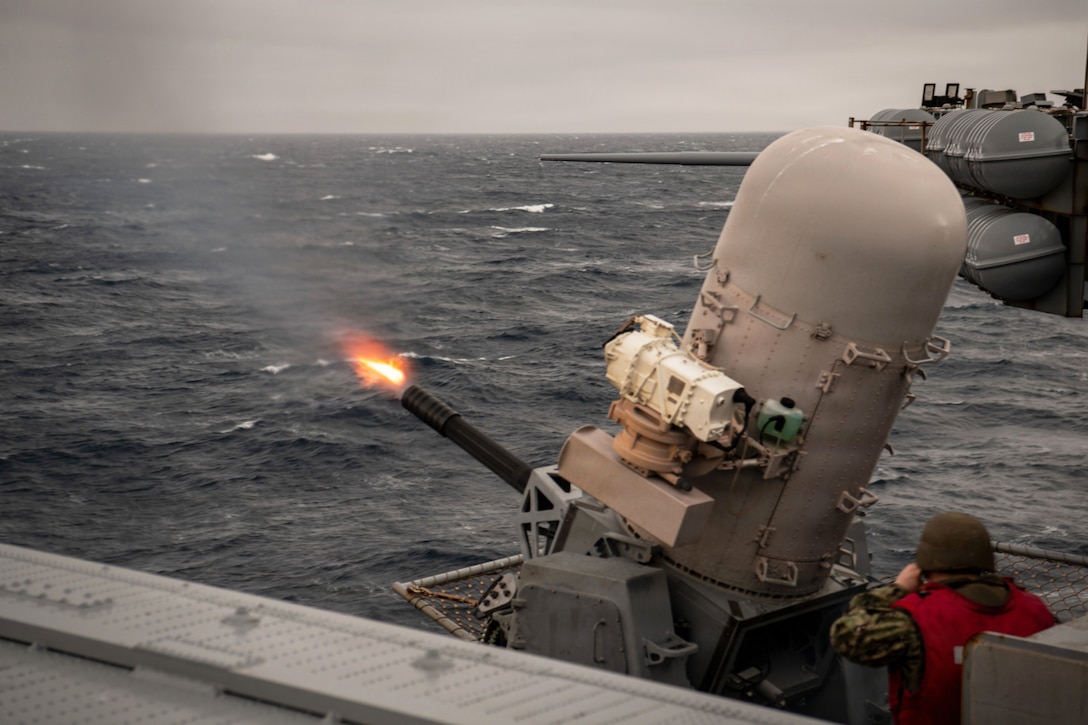 A sailor fires a weapon off the side of a ship at sea, creating a small orange flame on a gloomy day.