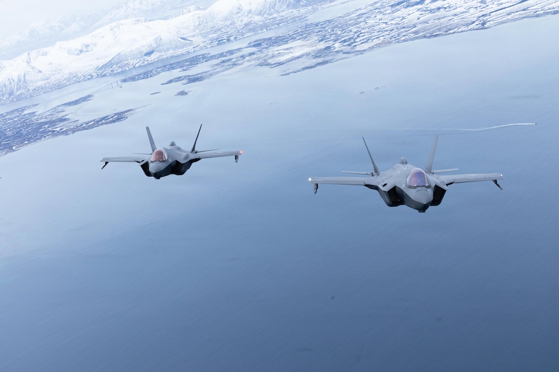 Two aircraft fly in formation over a body of water with snowy mountains in the background.