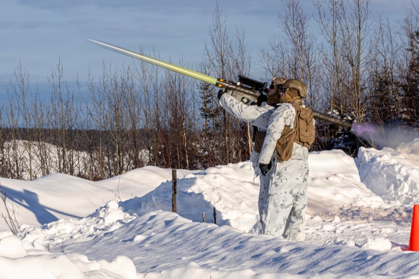 A soldier stands by as a fellow soldier, both in winter gear, fires a missile in a snowy field during the day.