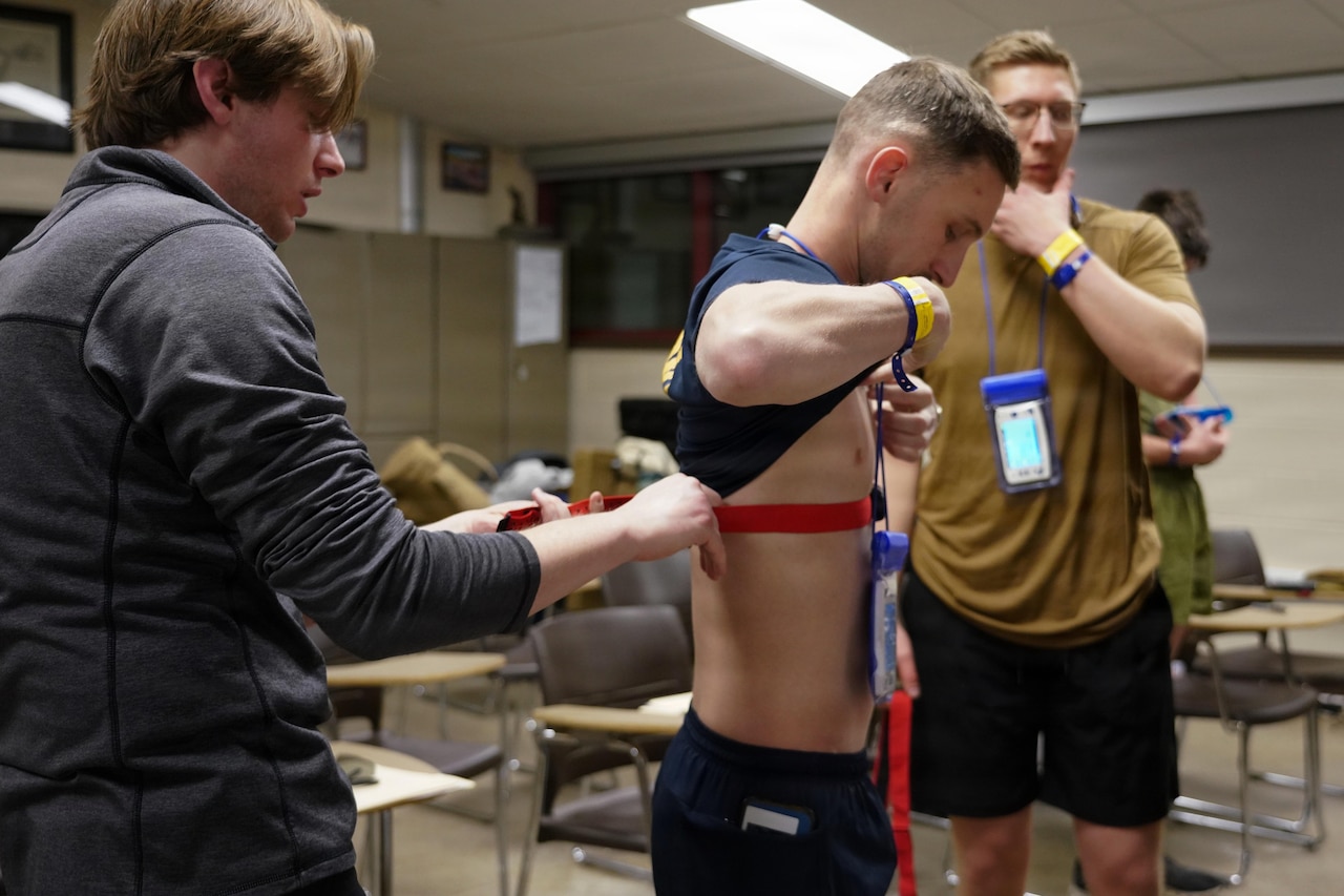 A man in casual attire attaches a medical device to the chest of another man, who is holding his shirt up while standing in a classroom. There is another man in athletic attire standing in the background.