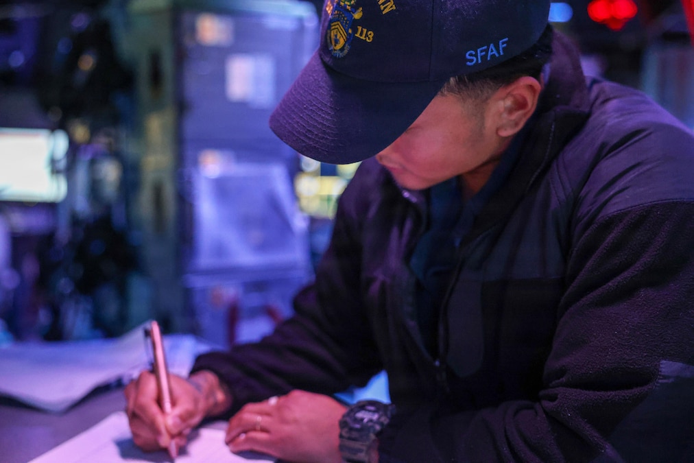 A sailor writes on a piece of paper in a dimly lit combat information center.