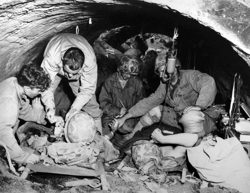 Four men stand or kneel above two men on stretchers, giving them aid. All are in a small concrete shelter not high enough to stand up in.