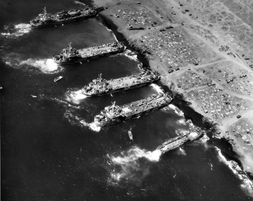 An aerial view of four large ships beached along a shoreline to deliver supplies.