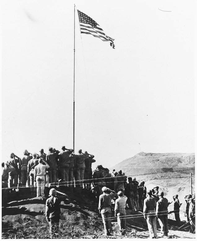 Dozens of men stand on a small, desolate hill to salute an American flag that’s been raised on a tall flagpole.