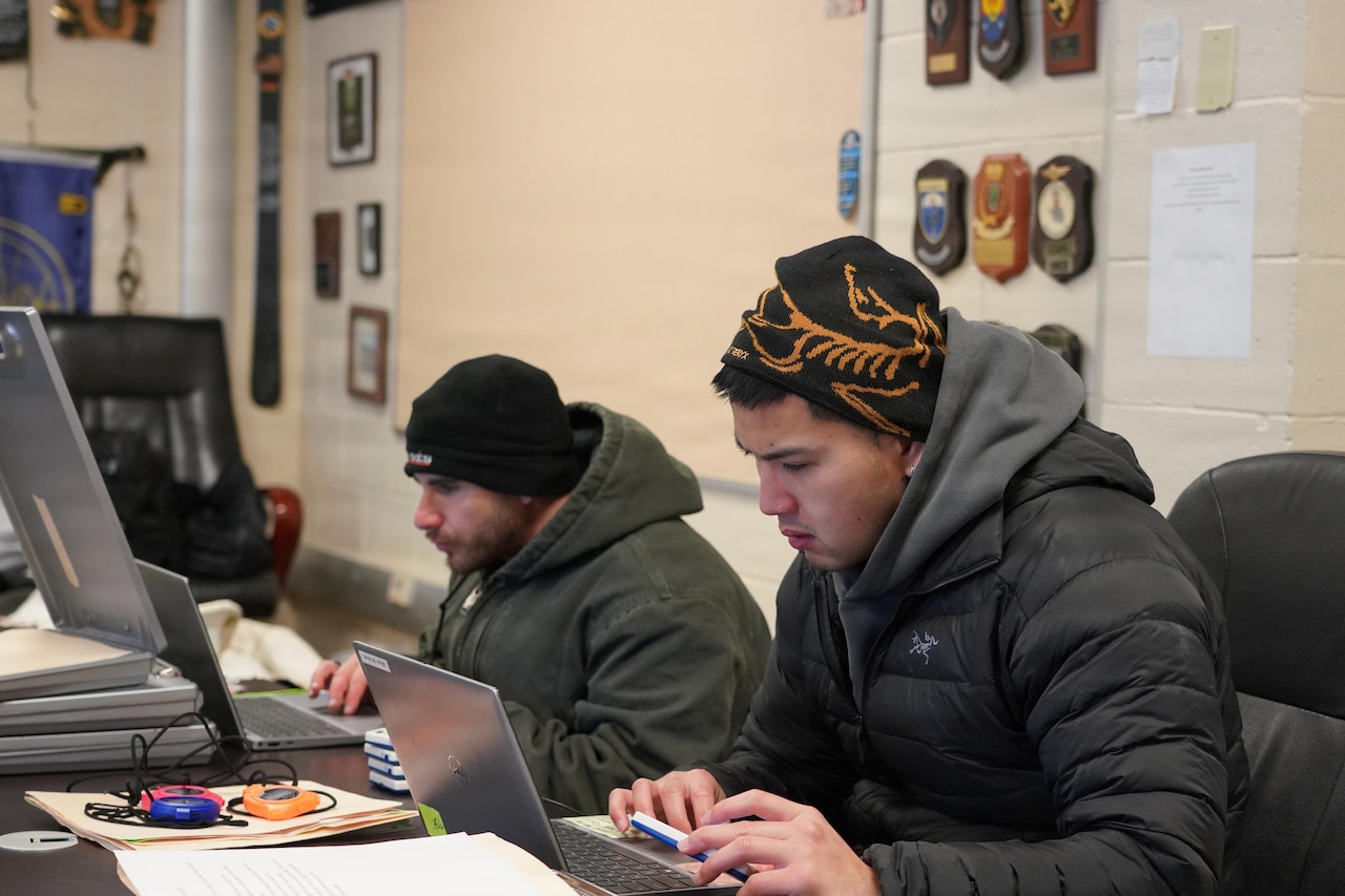 Two men in winter attire fill out forms on computers in a room. There are military awards on the walls in the background.