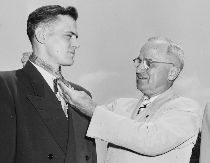 A man in business attire puts a medal around the neck of another man in similar attire, as two men in formal military uniforms observe.