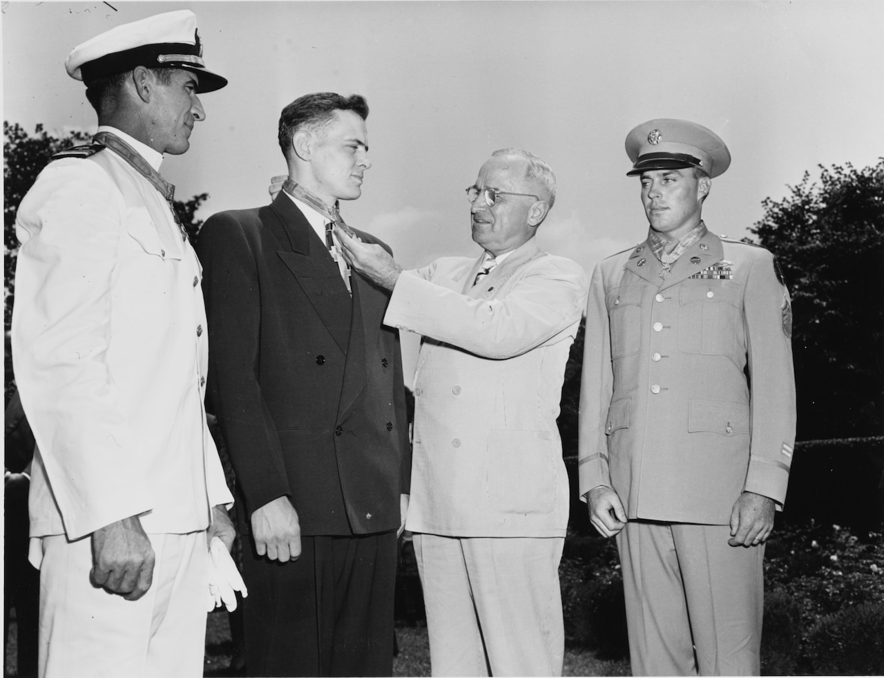 Three men in formal military uniforms stand together, with a man in business attire standing in the center, who places a medal around the neck of the man in the middle.