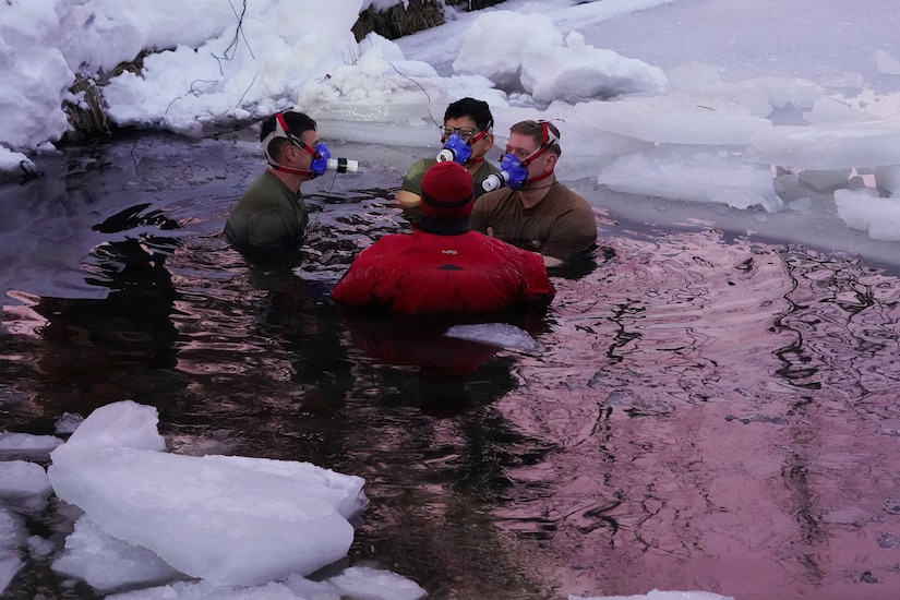 Three men wearing athletic attire and respirators stand in chest-deep cold water with chunks of ice around them. Another person in a cold-weather suit is standing in the water next to them.