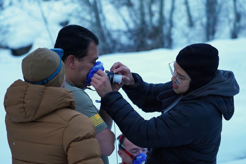 A man in winter attire places a medical device over the mouth of another man in athletic attire who is standing outside with snow on the ground. A third person in winter attire is standing next to the man in athletic attire.