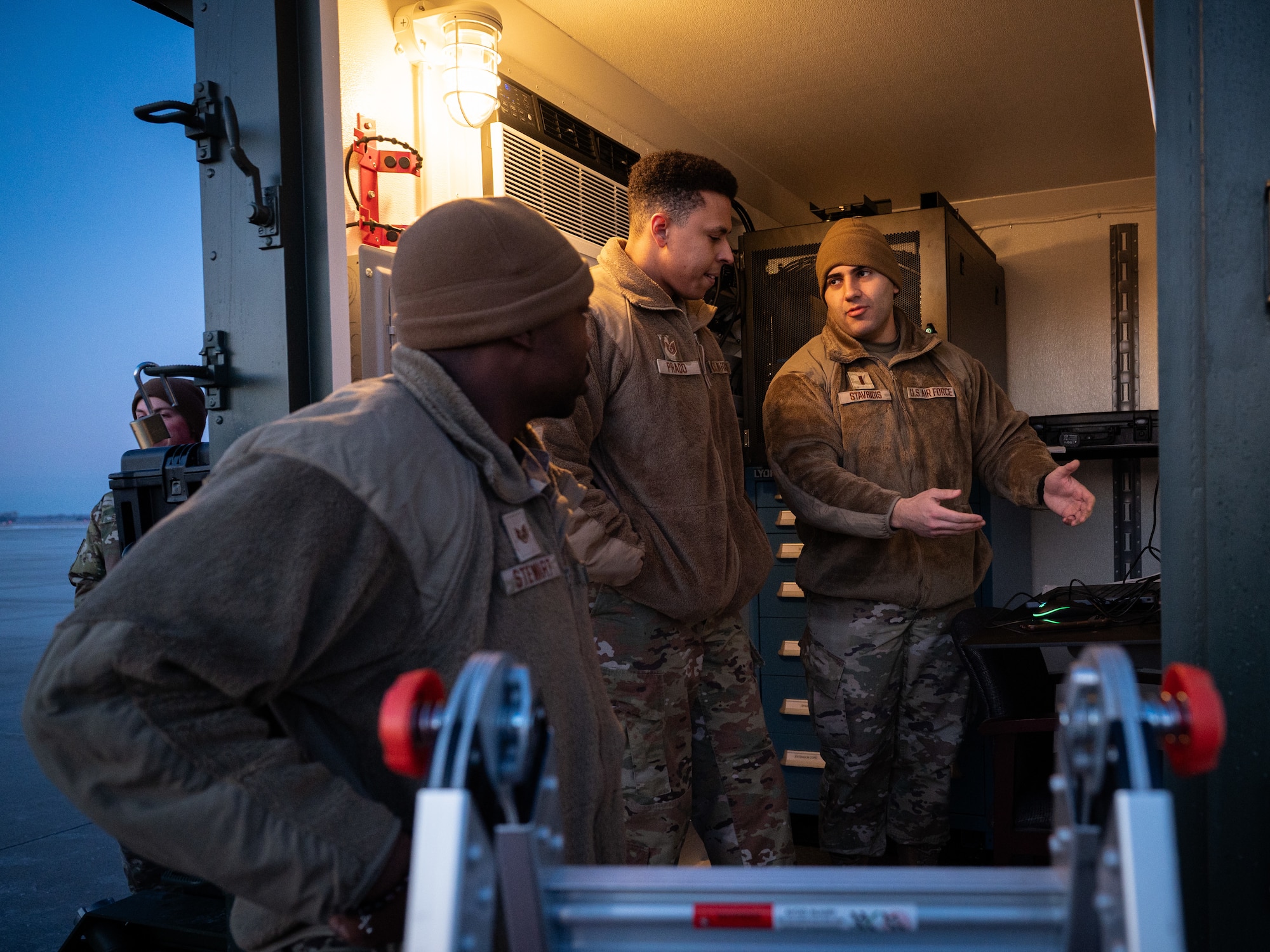 A photo of three men standing in a small room on a flightline.