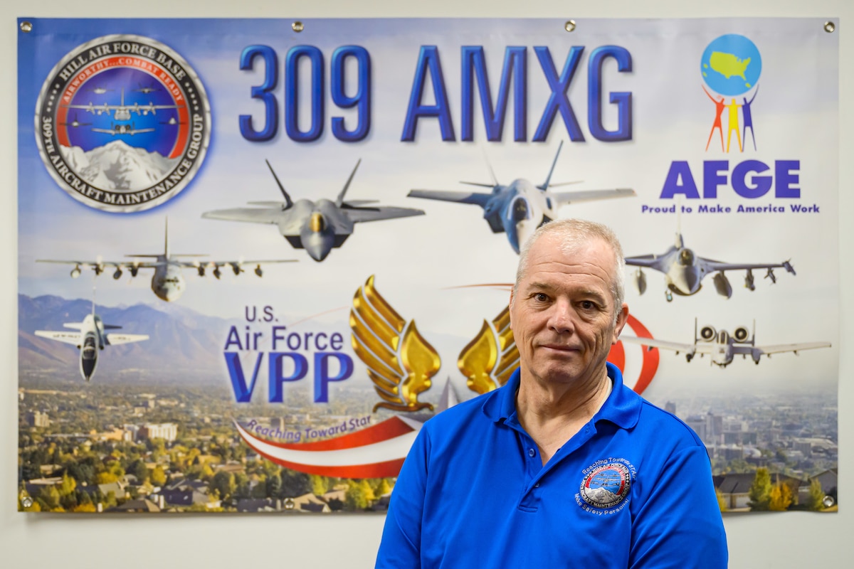 Edwards Takacs stands in front of a U.S. Air Force VPP poster displaying the 309th Aircraft Maintenance Group (309th AMXG) and images of the aircraft supported by the unit.