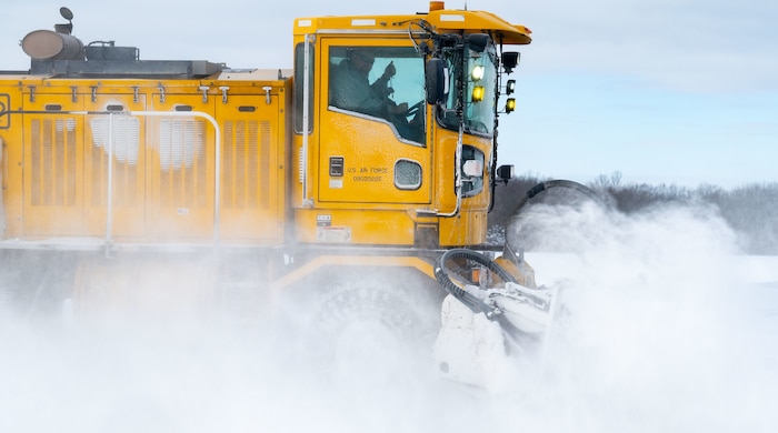 A man waves while driving a large yellow snowplow on the flightline