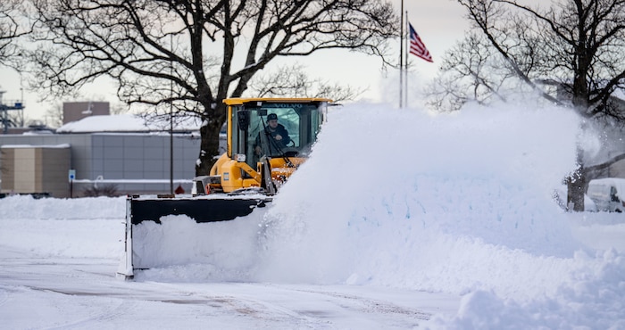 A man driving a yellow snowplow pushes a pile of snow in a parking lot.