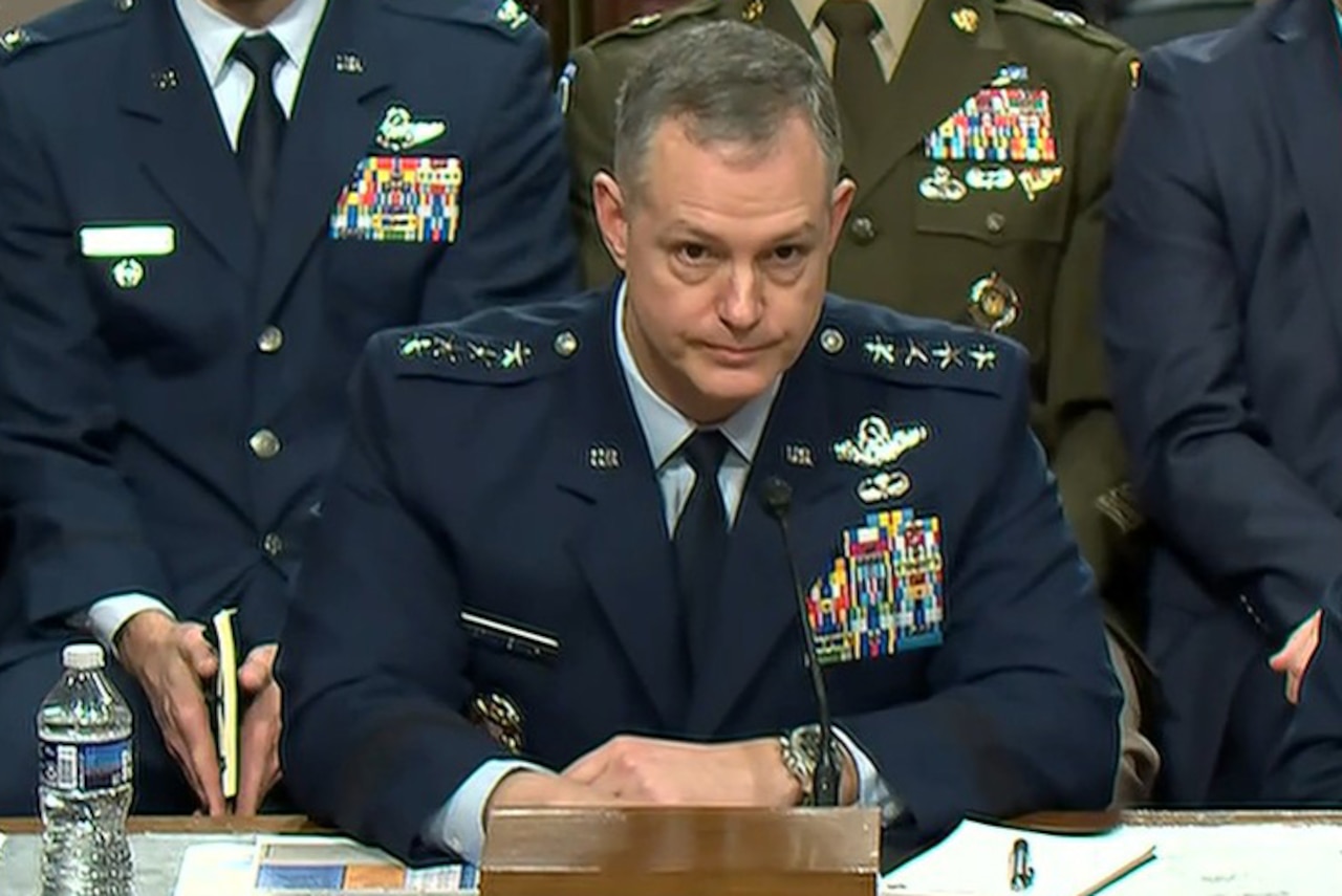 An airman speaks into a microphone while sitting at a table with people sitting in the background.