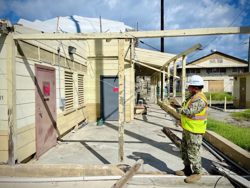 A man wearing a hard hat, dressed in a military camouflaged uniform and safety vest, stands near the partial shell of a building with a pen and pad in his hand.