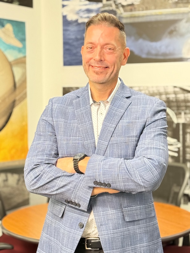 A man in a blue blazer smiles and stands with his arms crossed