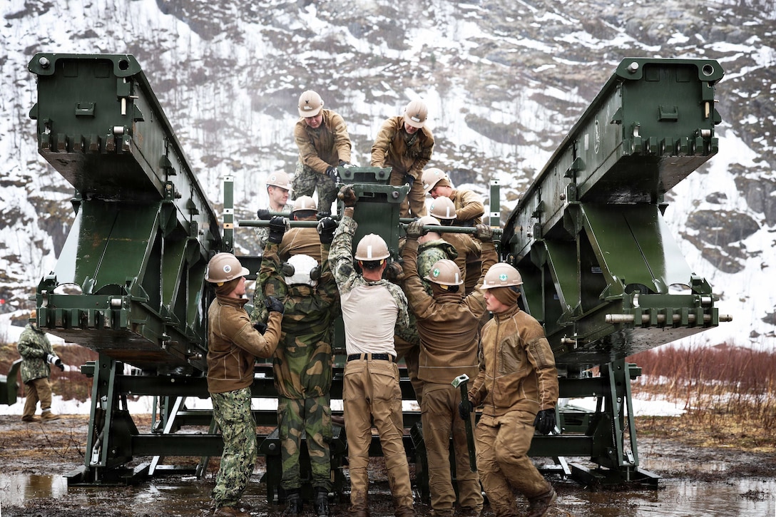 Sailors in construction gear work on a small green bridge in front of a snowy mountain.