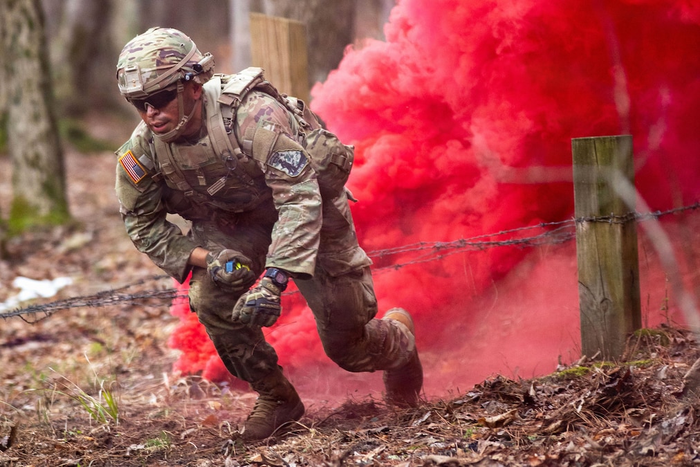 A soldier in tactical gear runs near a barbed wire obstacle course in a wooded area as a cloud of red smoke fills the background.