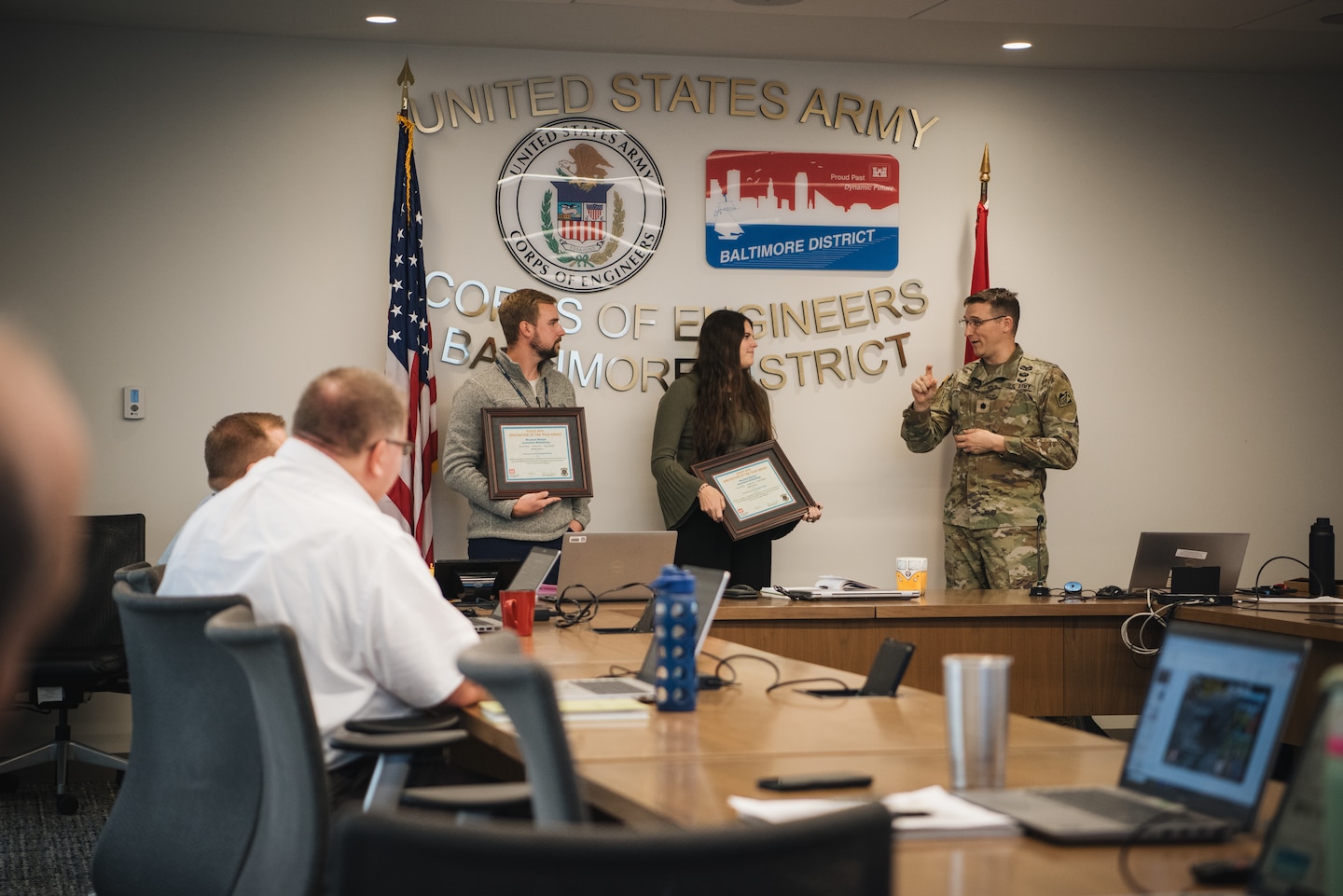 A uniformed Army officer speaks to a man and woman holding framed award certificates in a conference room. The wall behind them features the U.S. Army Corps of Engineers Baltimore District seal. People are seated at a table in the foreground, watching the presentation.