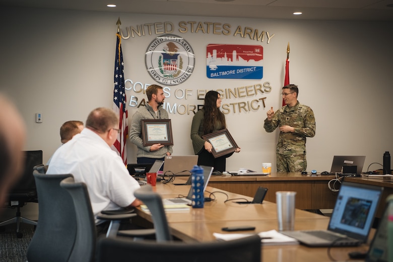 A uniformed Army officer speaks to a man and woman holding framed award certificates in a conference room. The wall behind them features the U.S. Army Corps of Engineers Baltimore District seal. People are seated at a table in the foreground, watching the presentation.