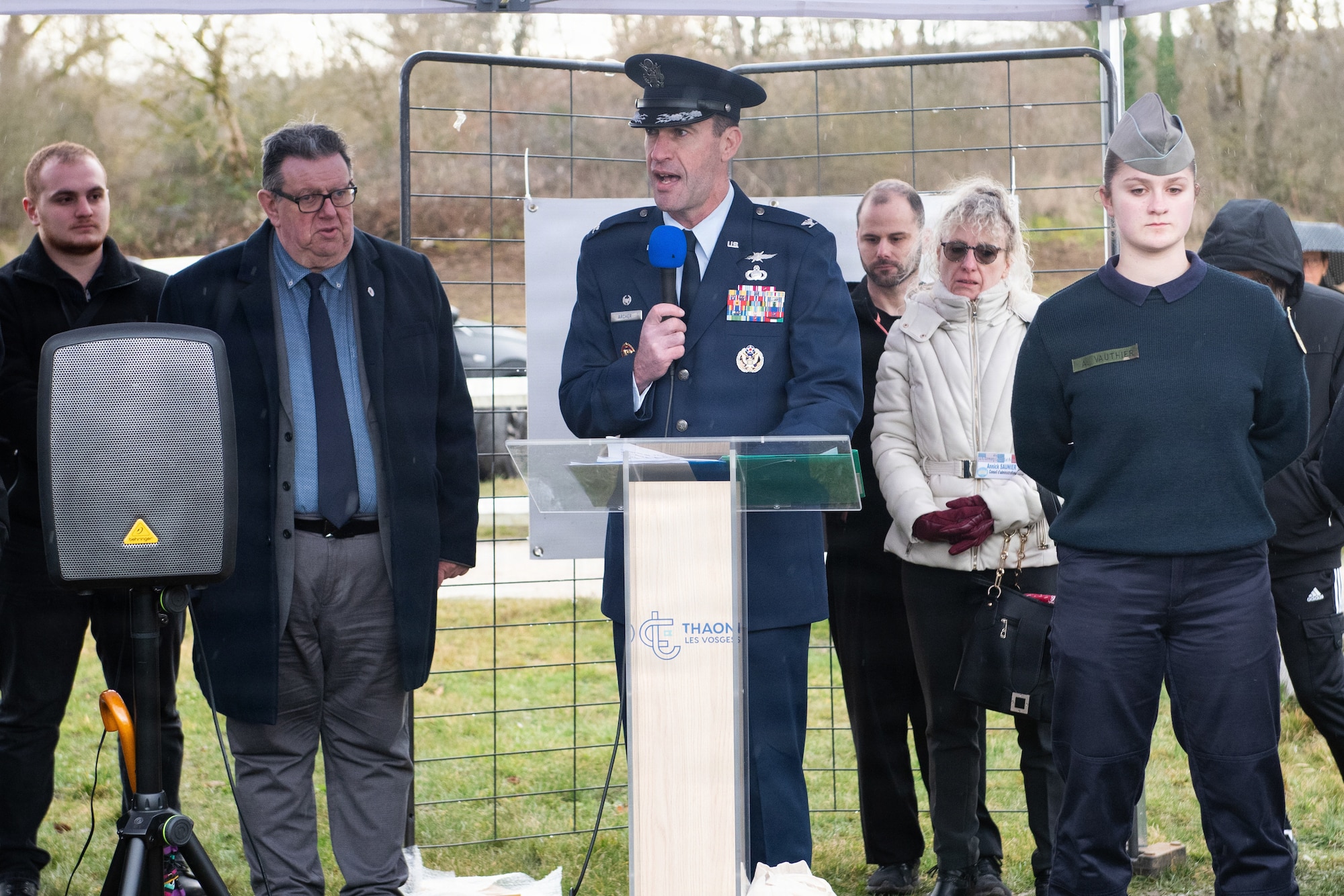 Photo shows an U.S. Air Force colonel flanked by a number of French residents, as he speaks at a podium during a memorial ceremony.