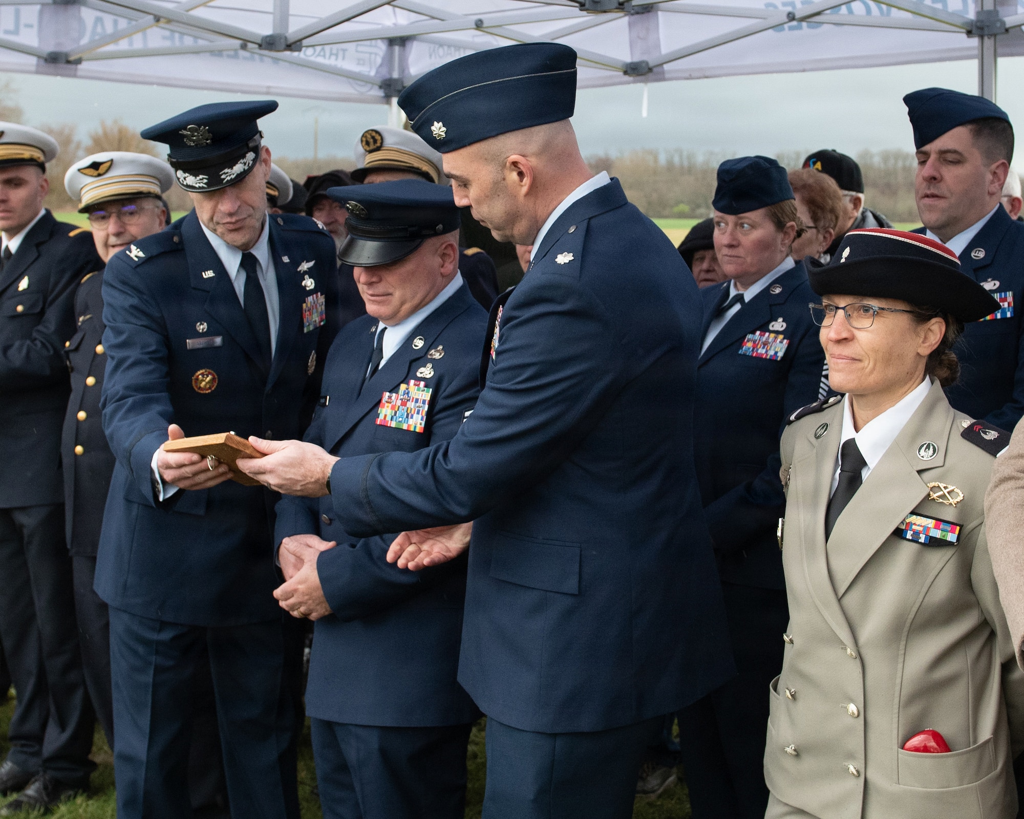 Photo shows several French residents and U.S. Airmen gathered outdoors for a memorial ceremony. Three Airmen are looking closely at a commemorative plaque.