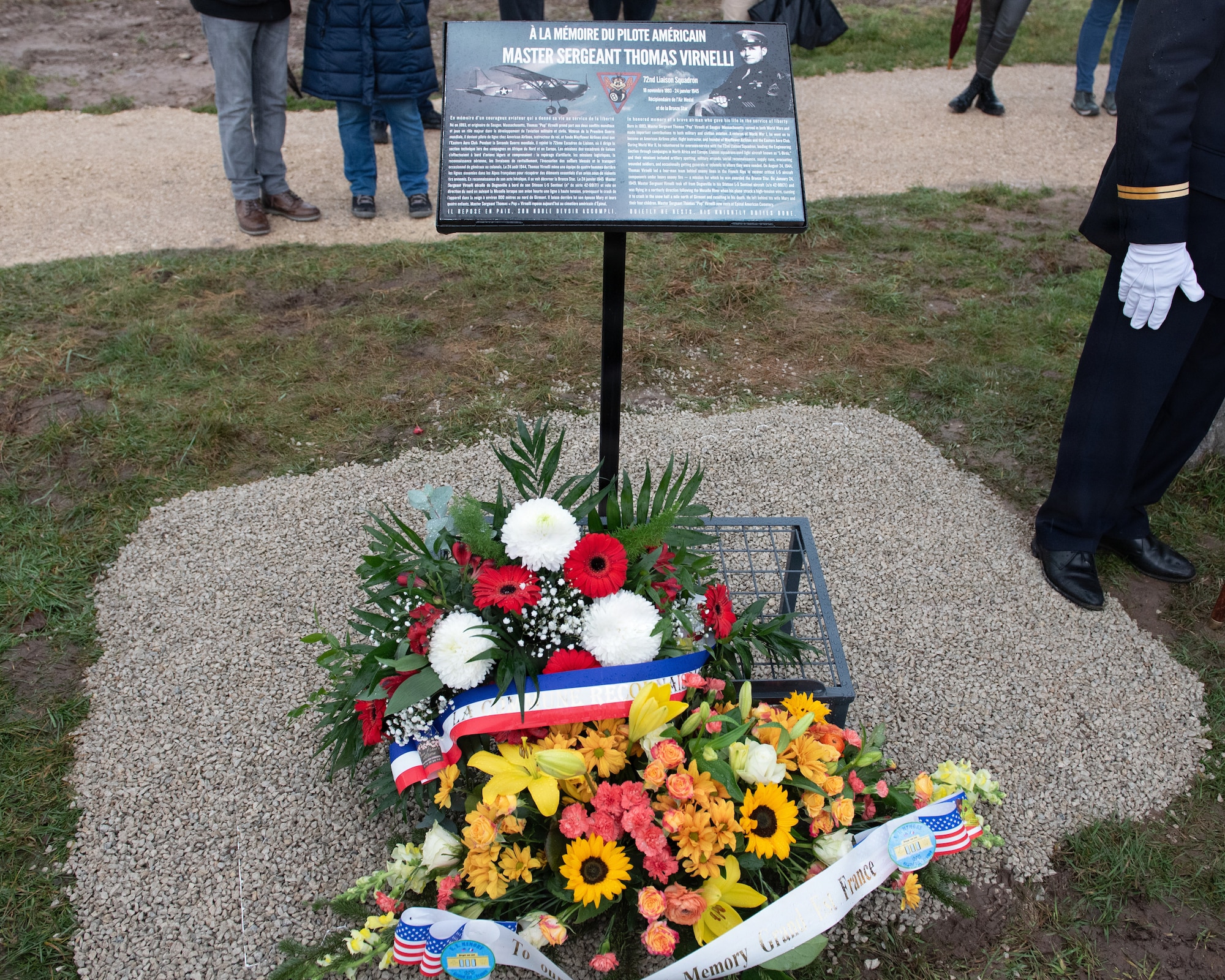 Photo shows a memorial plaque adorned by a floral bouquet during a ceremony.