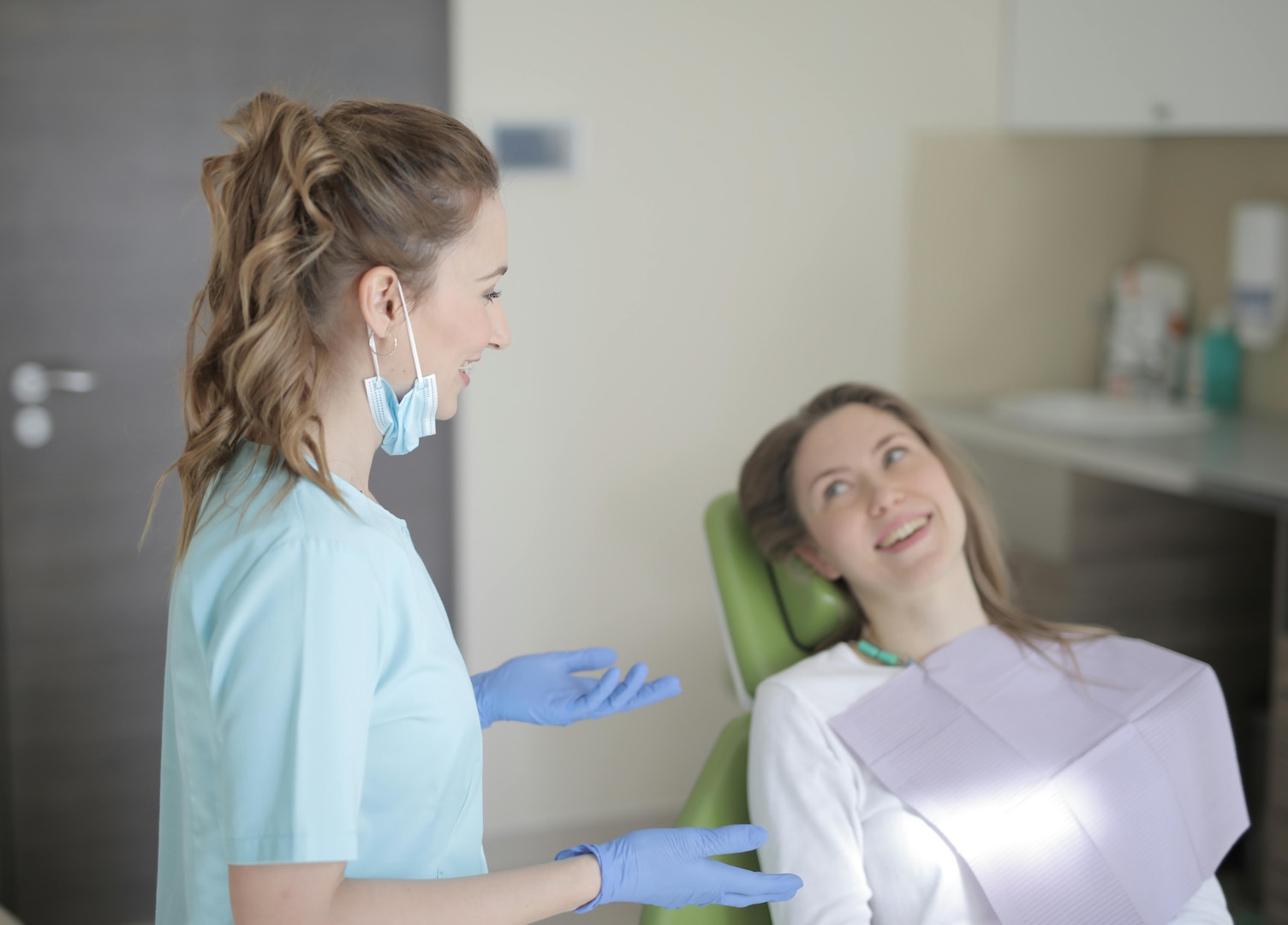 Woman in dentist's chair smiles up at dentist who's wearing scrubs and gloves.