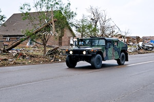 Humvee travels down road with tornado damage in background