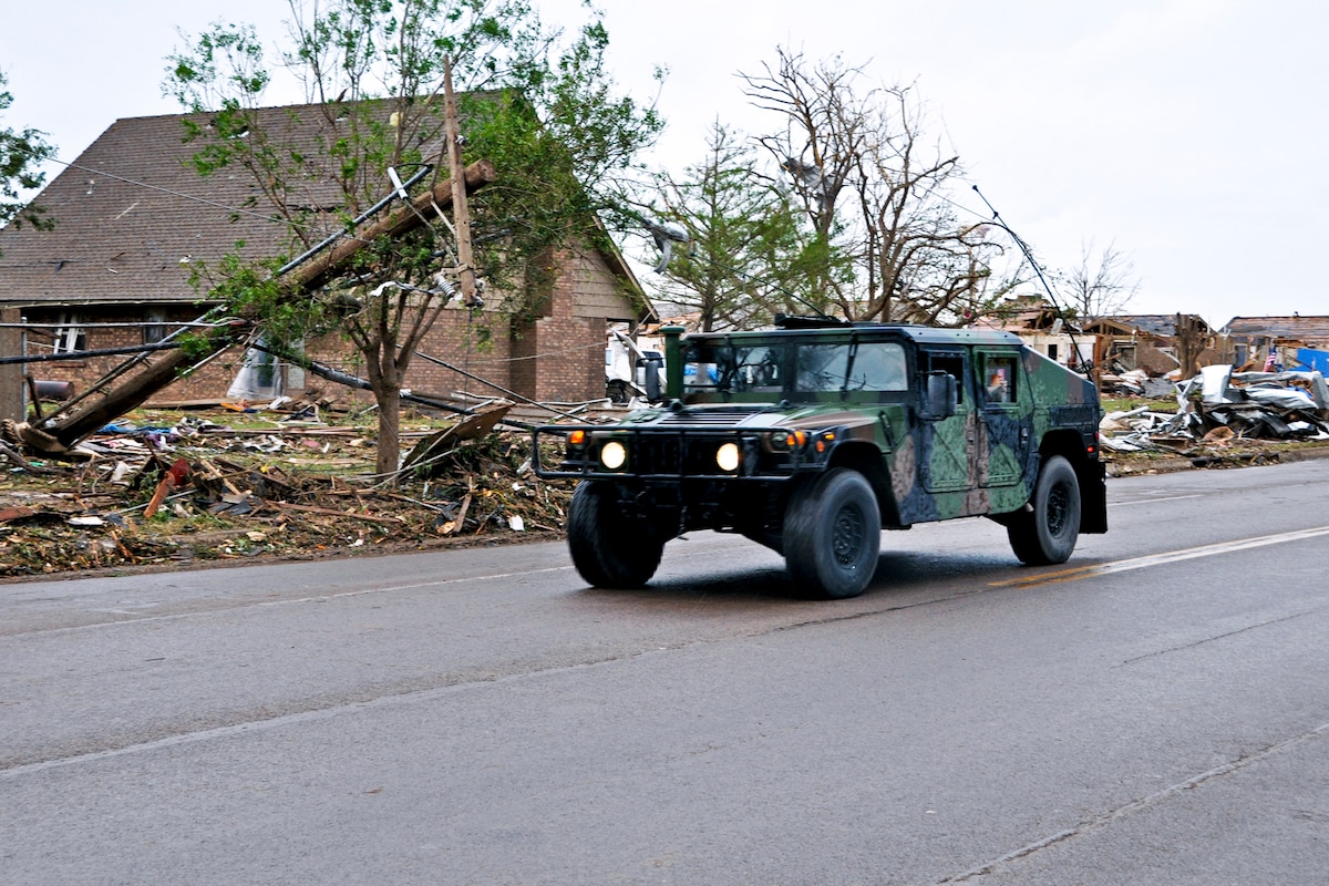 Humvee travels down road with tornado damage in background