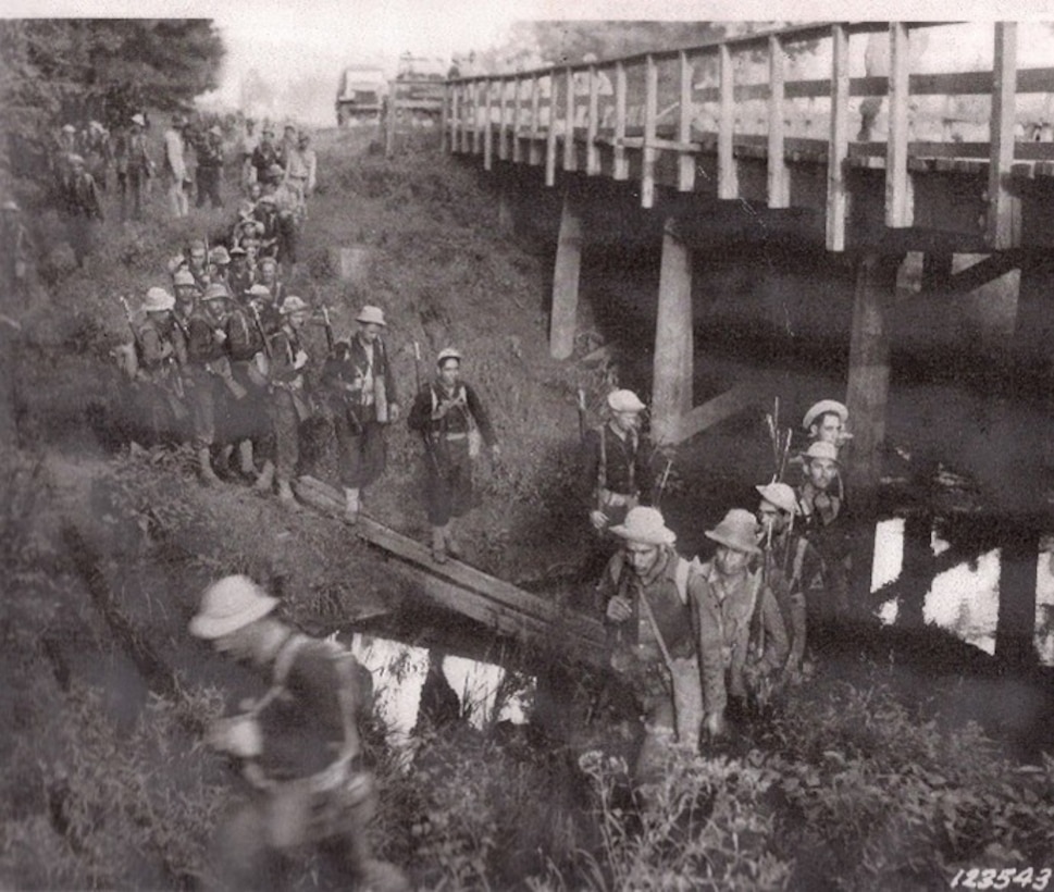 A line of soldiers cross a makeshift bridge across a creek