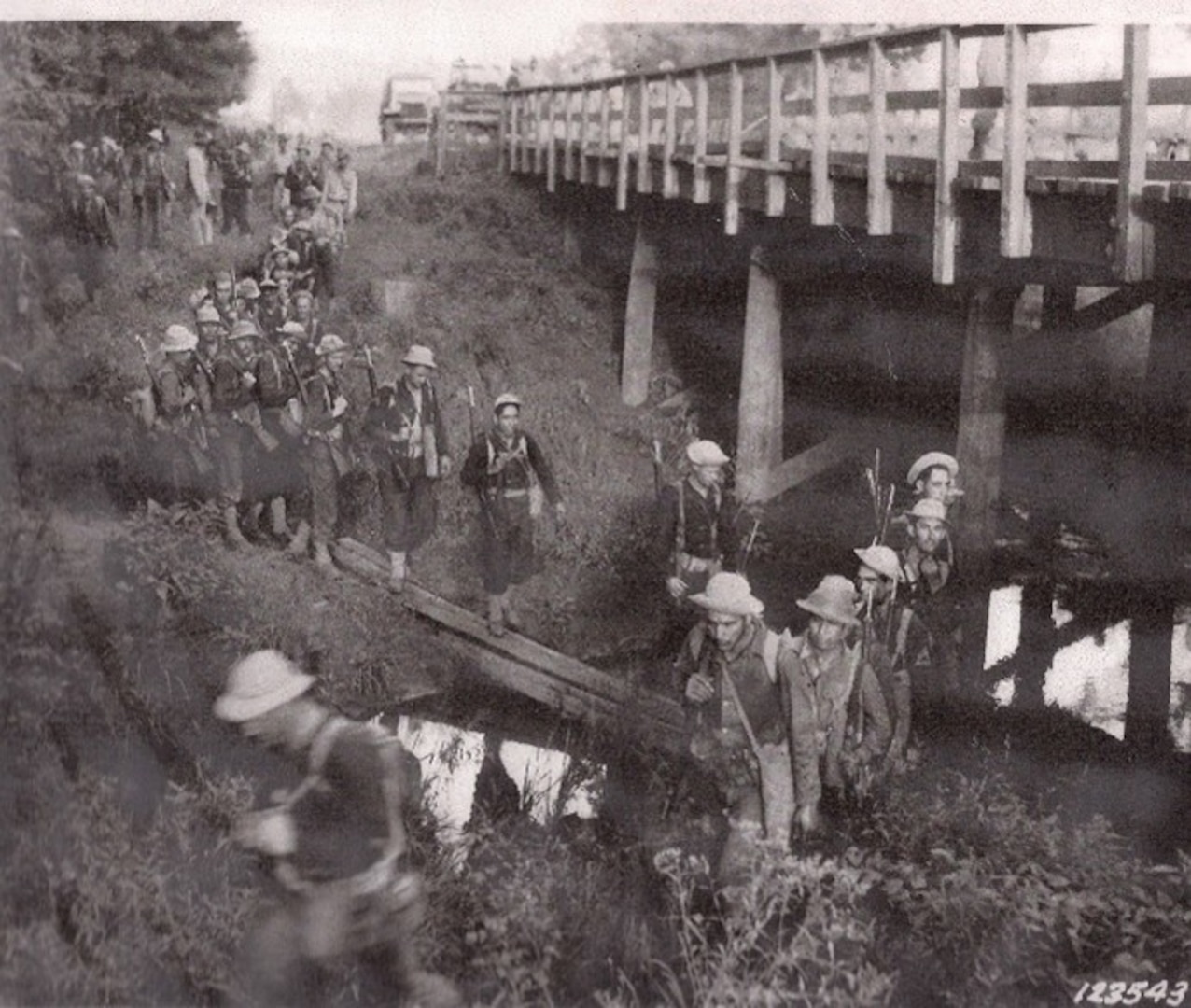 A line of soldiers cross a makeshift bridge across a creek