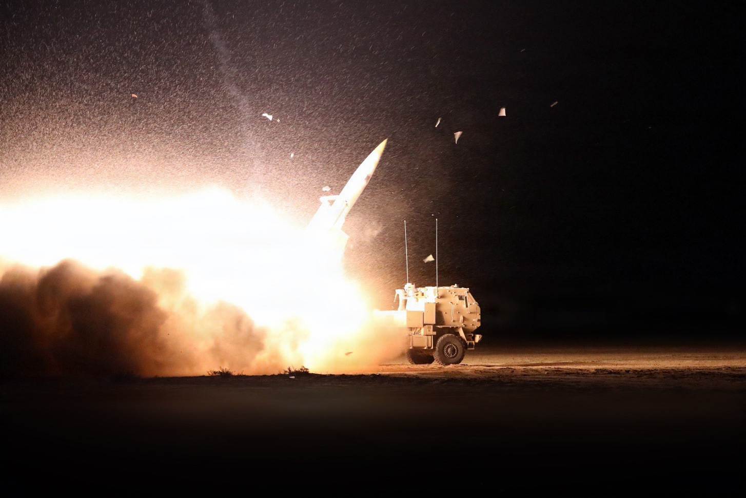 A missile launches from a military vehicle, creating a ball of fire and smoke against a dark sky.