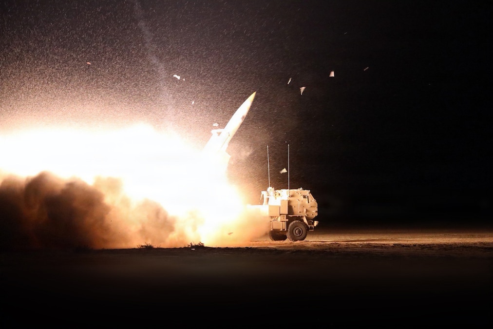 A missile launches from a military vehicle, creating a ball of fire and smoke against a dark sky.