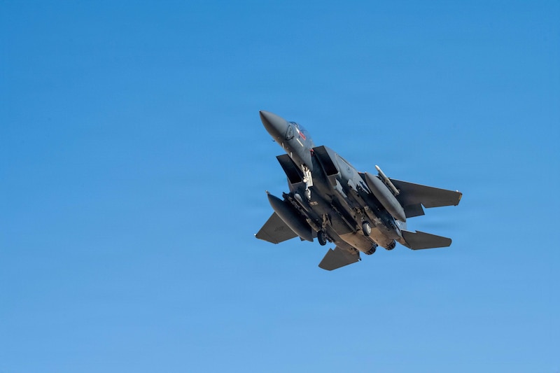 A military fighter jet with its landing gear deployed is flying under a clear daylight sky. 