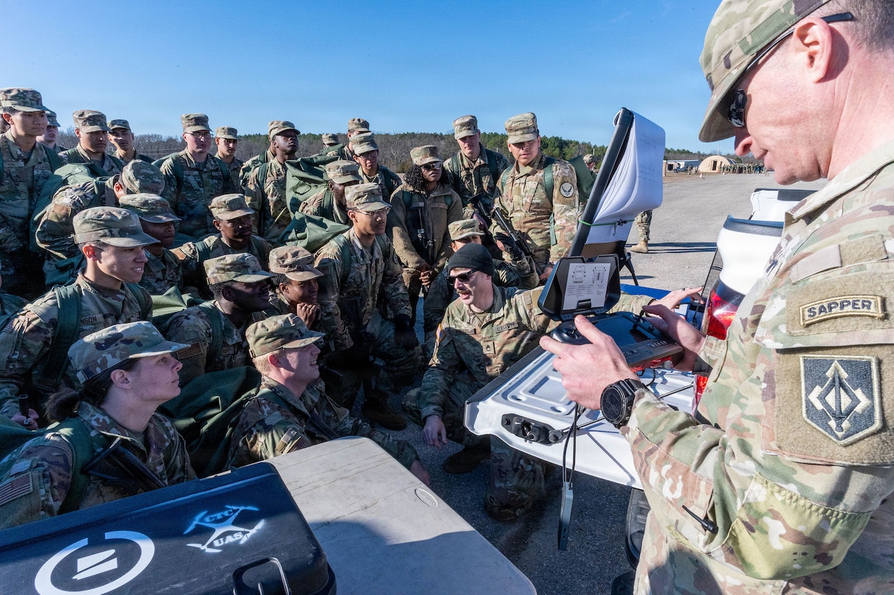 A man in a camouflage military uniform uses a remote control to pilot a drone while another man in similar attire crouches as he speaks to a group of people in camouflage military uniforms gathered outside.