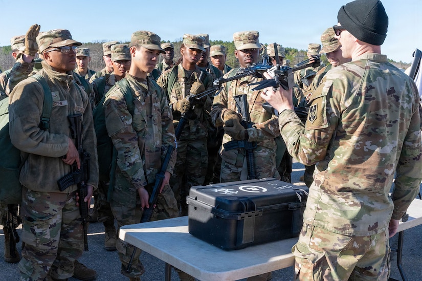 A man in a camouflage military uniform holds a drone while speaking to a group of people in similar attire gathered outside. In front of the man is a black case sitting on a table.