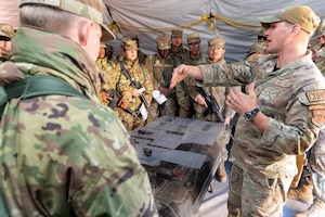 A man in a camouflage military uniform speaks to a group of people in similar attire holding rifles while they stand in a tent.