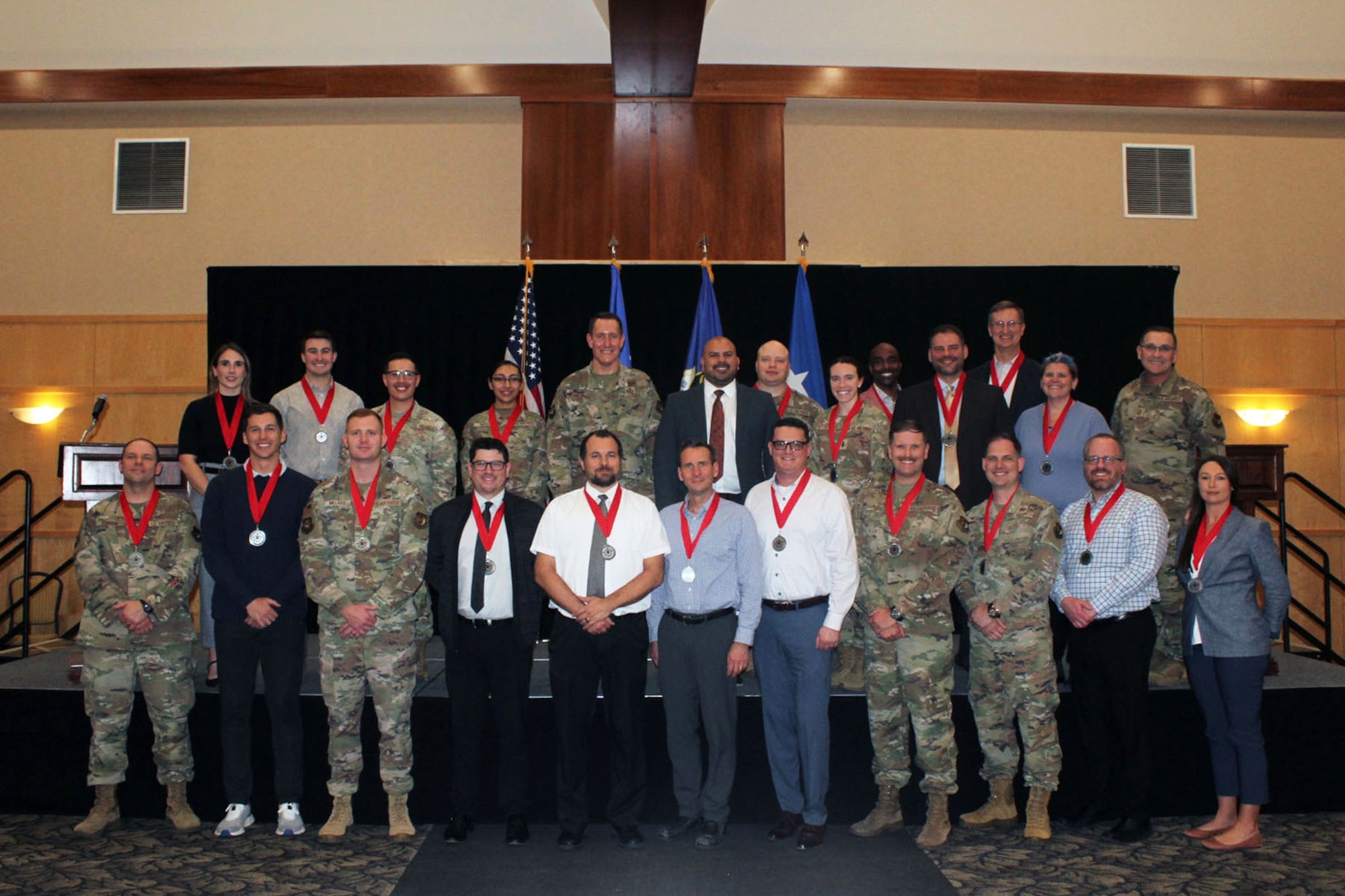 A group of men and women in civilian clothes and military uniforms stand in front of a stage wearing medals