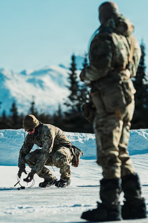 A man wearing a winter camouflage military uniform lays wire onto a snow-covered ground. Another man in similar attire stands and watches.