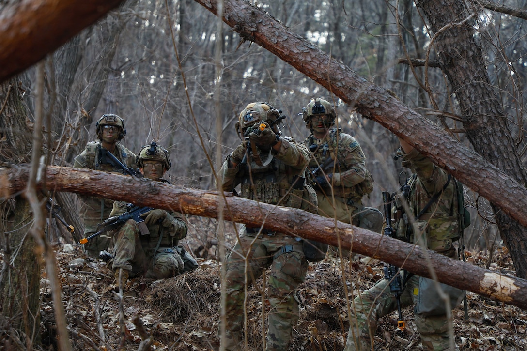 Two soldiers in camouflage lie next to each other in a field as one aims weapon loaded with ammo.