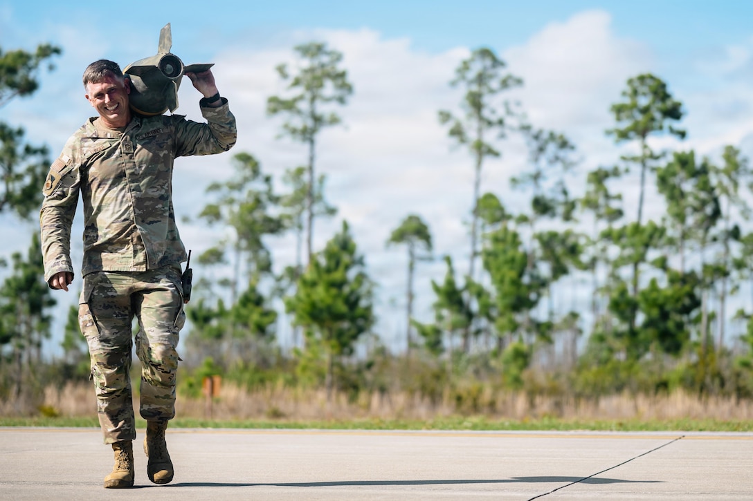 An airman carries ordnance over their left shoulder while walking in a parking lot under a blue sky.